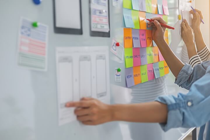 A picture of a white board with sticky notes and planning. People's hands are writing on the board