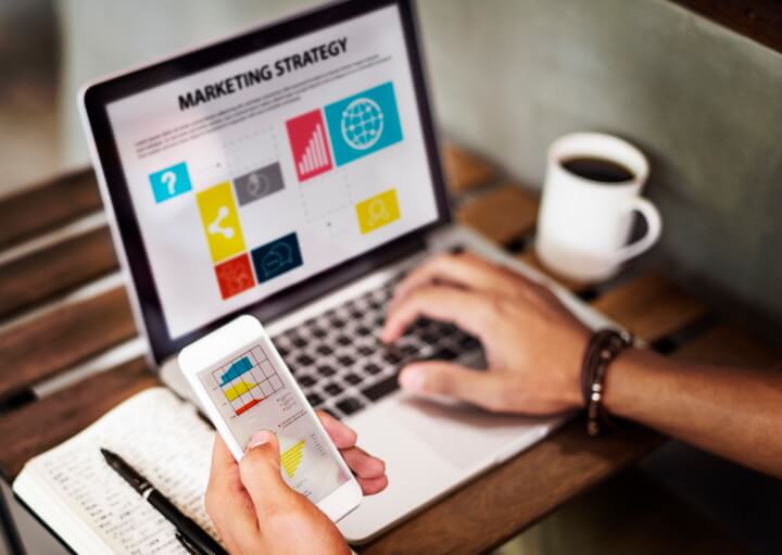 A picture of a man holding a smart phone with a laptop in the background.